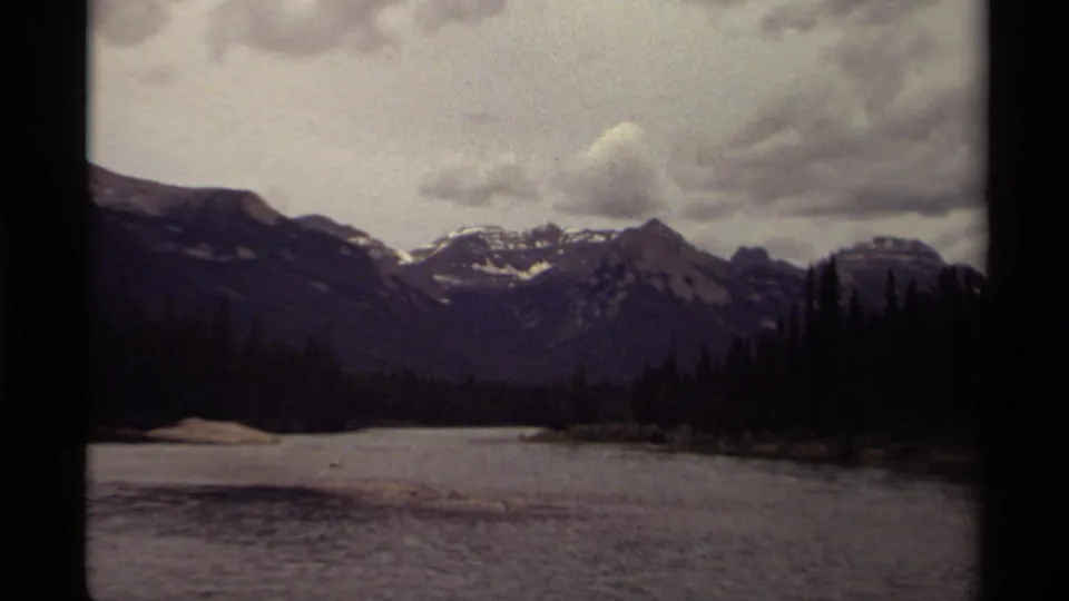 Canadian 1979 home-movie frame holding on a cloudy mountain and lake scene with dark trees at the shoreline.