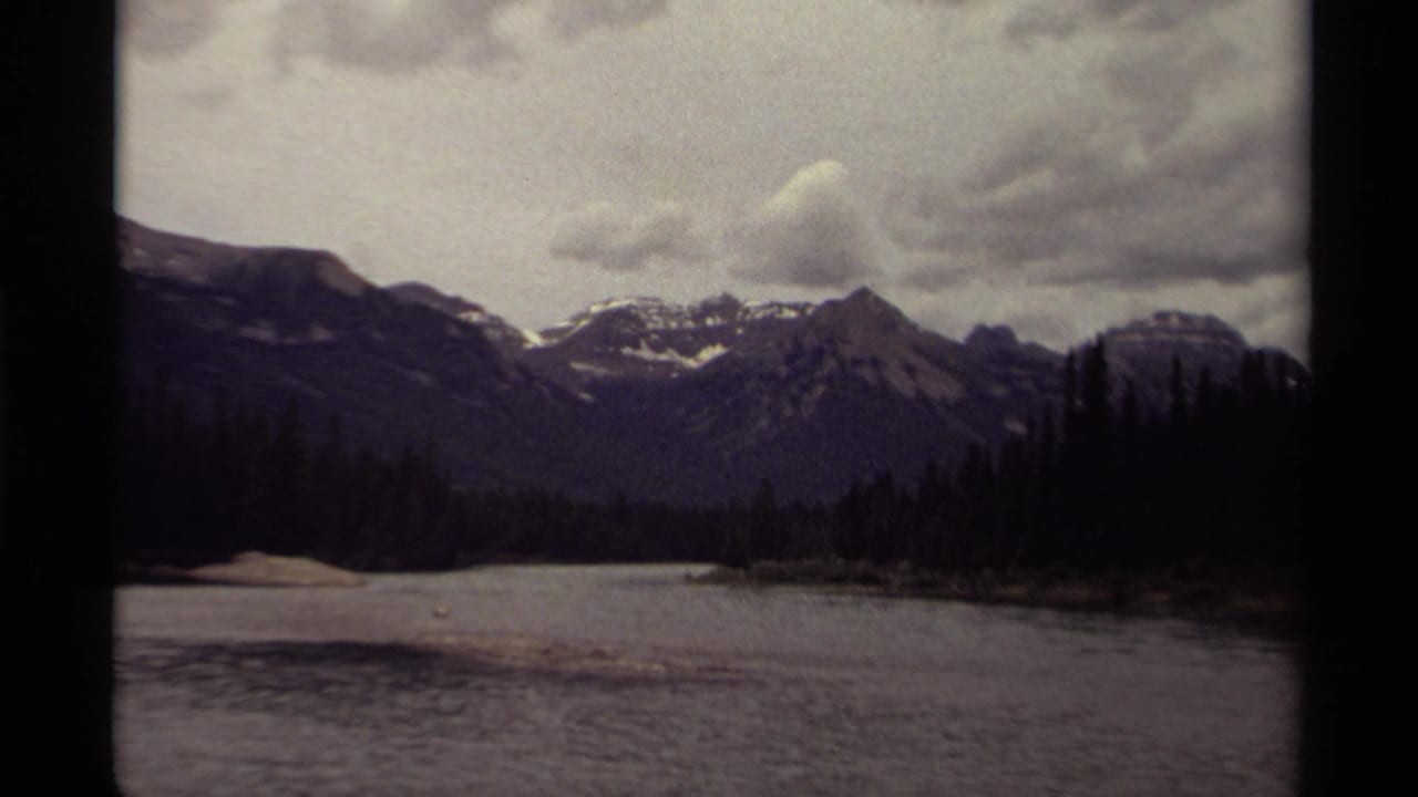 1979 Canadian 8mm view of snow-streaked mountains, evergreen shoreline, and still water beneath a gray overcast sky.