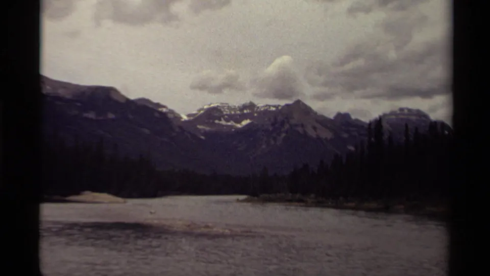 1979 Canadian 8mm view of snow-streaked mountains, evergreen shoreline, and still water beneath a gray overcast sky.