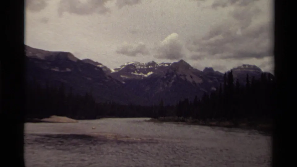 1979 Canadian wilderness scene with distant peaks, dense evergreens, and a quiet lake or river under cloud cover.
