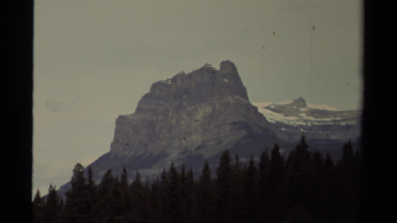 1979 Canadian 8mm frame showing a rugged mountain peak behind dark evergreen trees beneath a pale sky.
