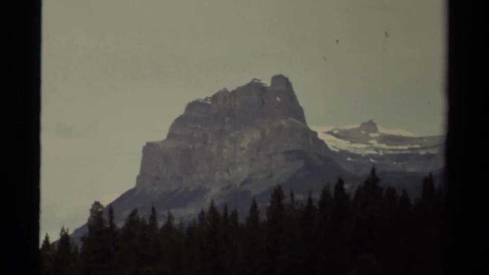 1979 Canadian 8mm frame showing a rugged mountain peak behind dark evergreen trees beneath a pale sky.