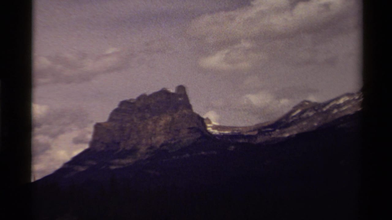 1979 Canadian home-movie frame with a rocky peak, snow-dusted ridge line, dark foreground slope, and cloudy sky.