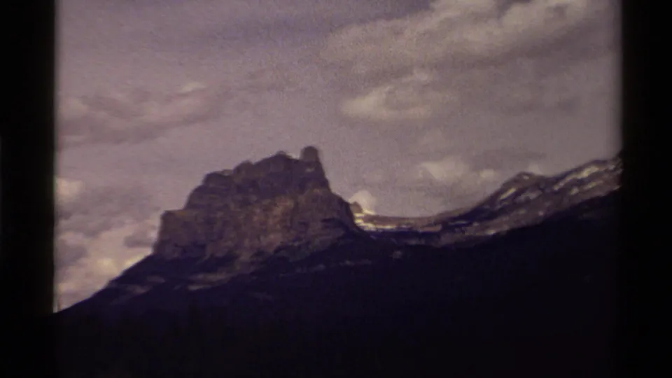 1979 Canadian home-movie frame with a rocky peak, snow-dusted ridge line, dark foreground slope, and cloudy sky.