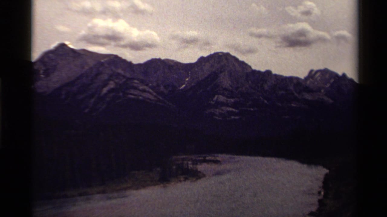1979 Canadian 8mm frame showing a snow-streaked mountain ridge, forested slopes and a curving waterway in the valley.