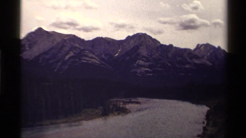 1979 Canadian home-movie frame of a forested mountain valley with a broad waterway and rugged peaks beneath scattered clouds.