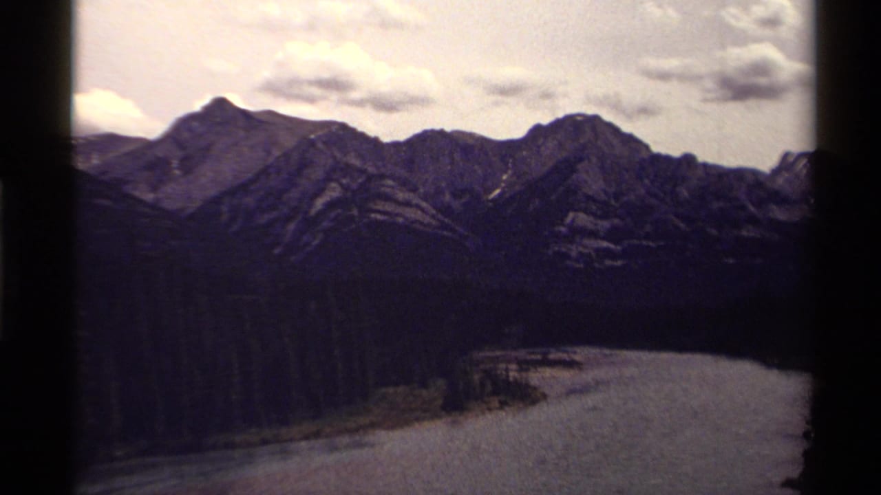 1979 Canadian 8mm frame showing rugged mountains, evergreen forest, and a broad river bend beneath scattered clouds.