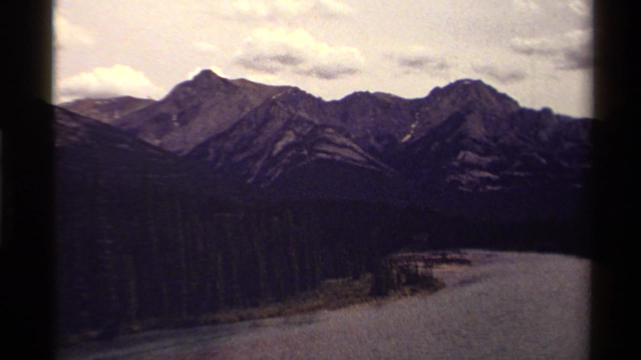 Canadian 8mm travel view of layered mountain peaks, dark evergreen forest, and water curving along the valley edge.