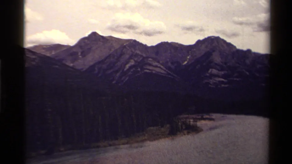 Canadian 8mm travel view of layered mountain peaks, dark evergreen forest, and water curving along the valley edge.
