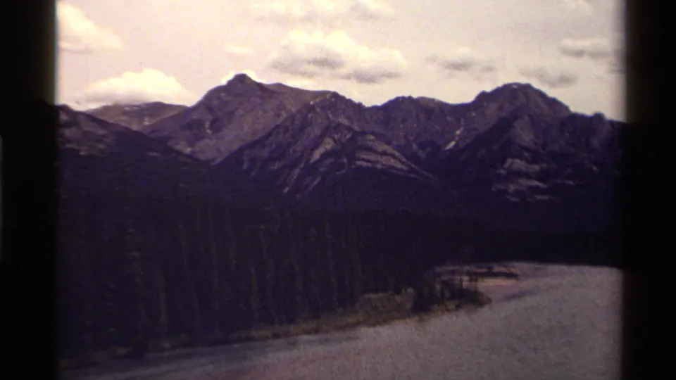 1979 Canadian 8mm frame with rocky peaks, dense forest, and a broad water surface at the bottom of the view.