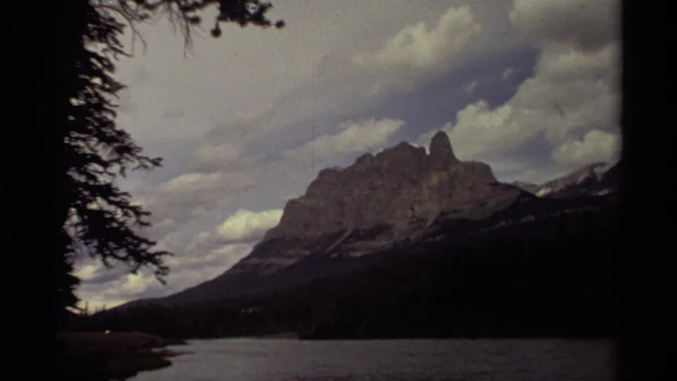 Canadian 1979 home-movie frame showing a mountain ridge over a lakeshore, partly framed by dark trees and overcast sky.