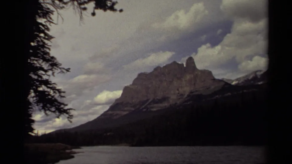 1979 Canadian 8mm frame of a mountain across a lake, with dark foreground trees and a cloudy sky in faded color.