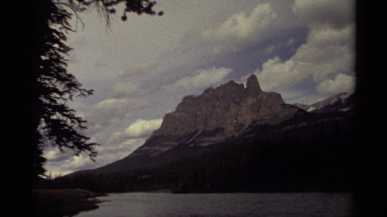 1979 Canadian 8mm frame showing a rugged mountain peak, dark forest, and lake foreground beneath layered clouds.
