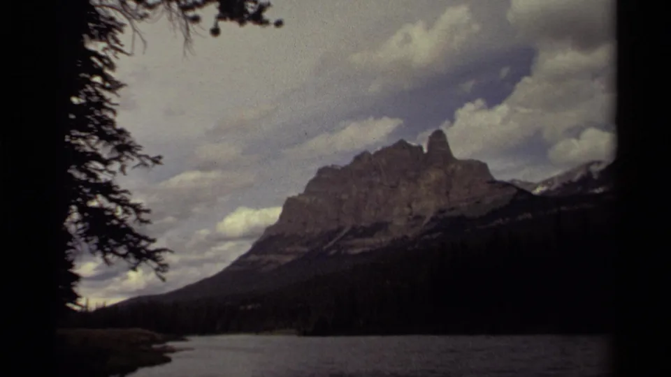 1979 Canadian 8mm frame showing a rugged mountain peak, dark forest, and lake foreground beneath layered clouds.
