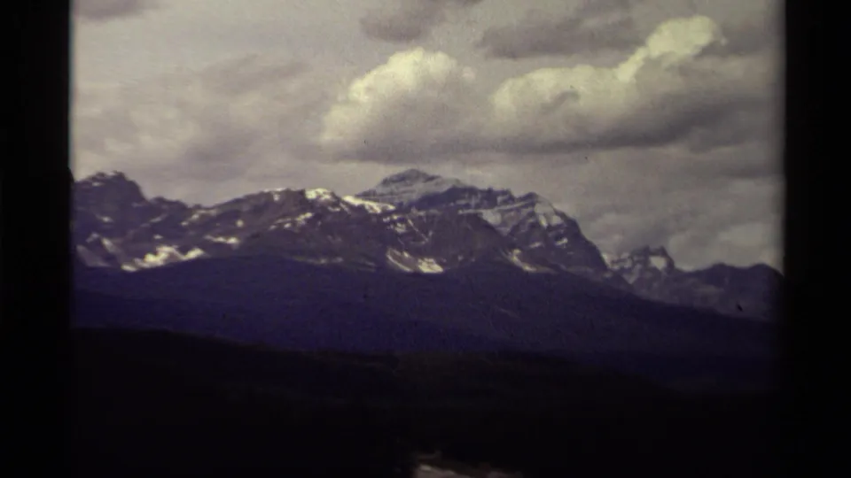 1979 Canadian 8mm frame showing a snow-patched mountain range over a dark forested valley beneath large clouds.
