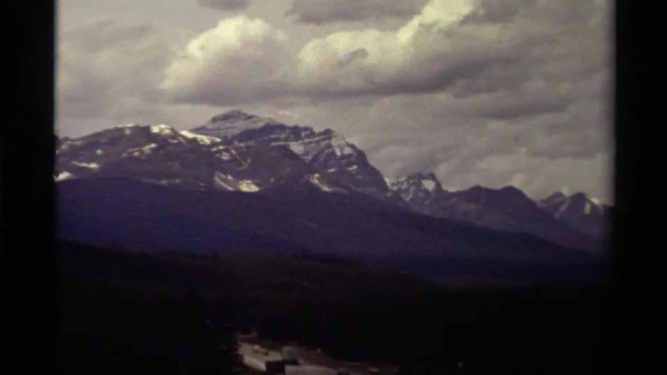 1979 Canadian 8mm frame with a snow-marked ridge, distant peaks, forested foreground, and cloud-filled sky.