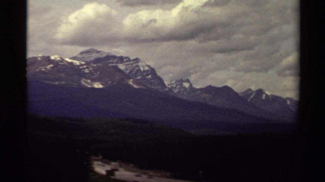 1979 Canadian 8mm frame of distant snow-streaked mountain peaks beneath layered twilight clouds and a dark foreground slope.