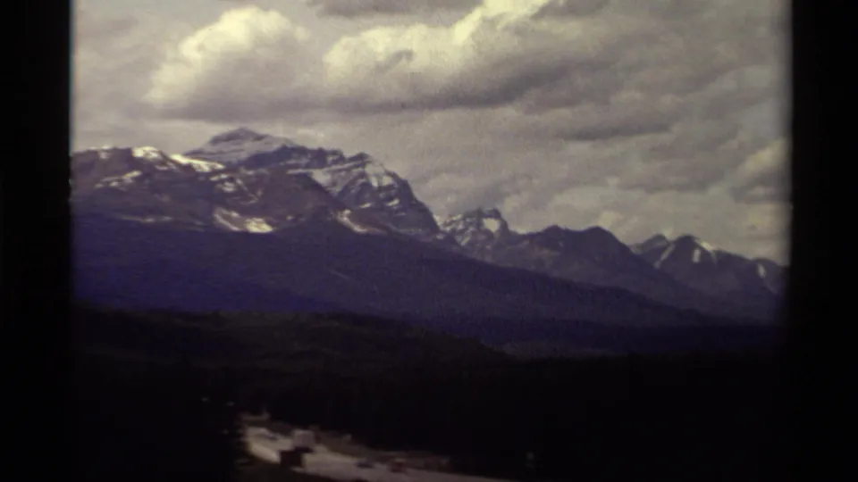 1979 Canadian 8mm frame of distant snow-streaked mountain peaks beneath layered twilight clouds and a dark foreground slope.