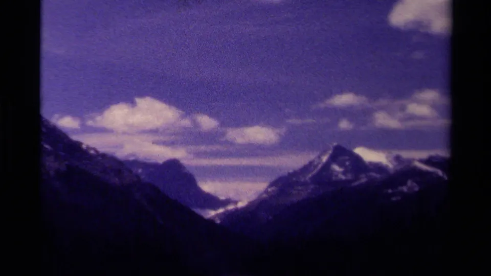 Wide 1979 Canadian 8mm view of a mountain valley and snow-capped peaks beneath scattered clouds, with dark slopes in the foreground.