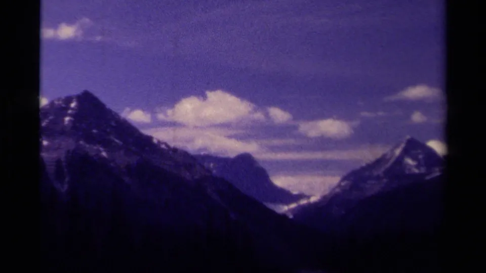 Vintage 8mm Canadian park landscape showing snow-capped peaks, a shadowed valley, and scattered clouds in a color-shifted sky.