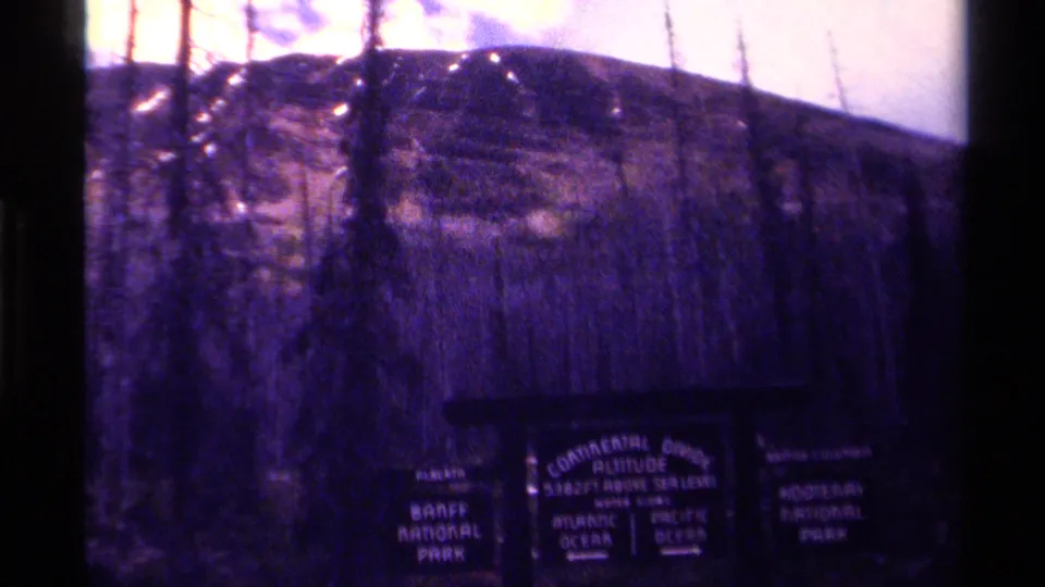 1979 Canadian 8mm frame of a wooden Continental Divide sign with Banff and Kootenay National Park names visible beneath bare trees.