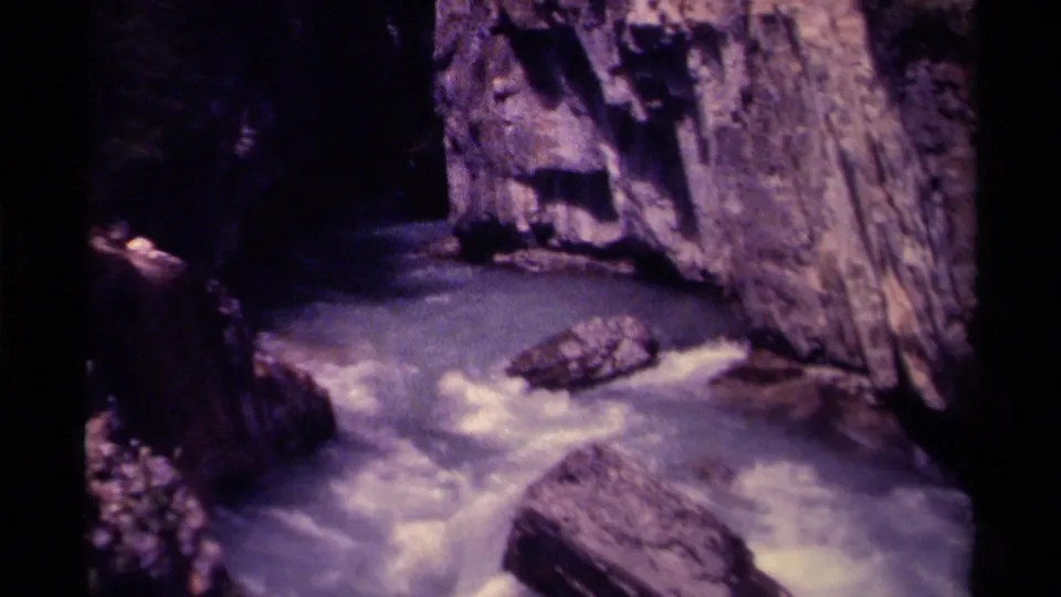 1979 Canadian 8mm frame of a rocky gorge where swift water turns around exposed boulders beneath high cliff faces.