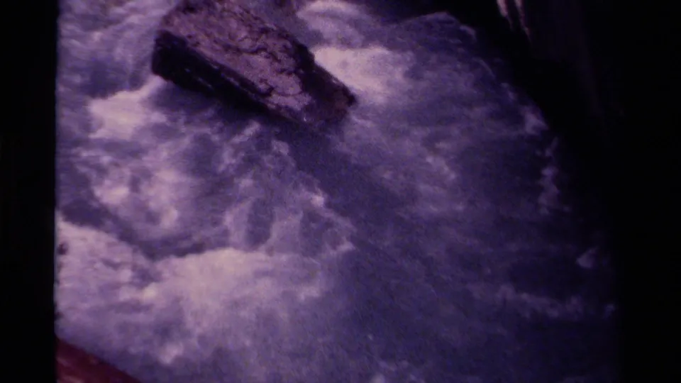 Handheld 8mm view of whitewater surrounding a prominent rock in a Canadian stream, with dark canyon borders and visible film grain.