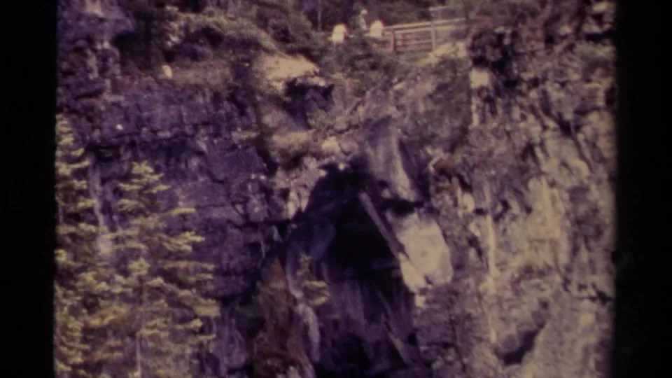 1979 Canadian 8mm frame showing a steep rock passage, evergreen growth, and a shadowed cave-like opening.