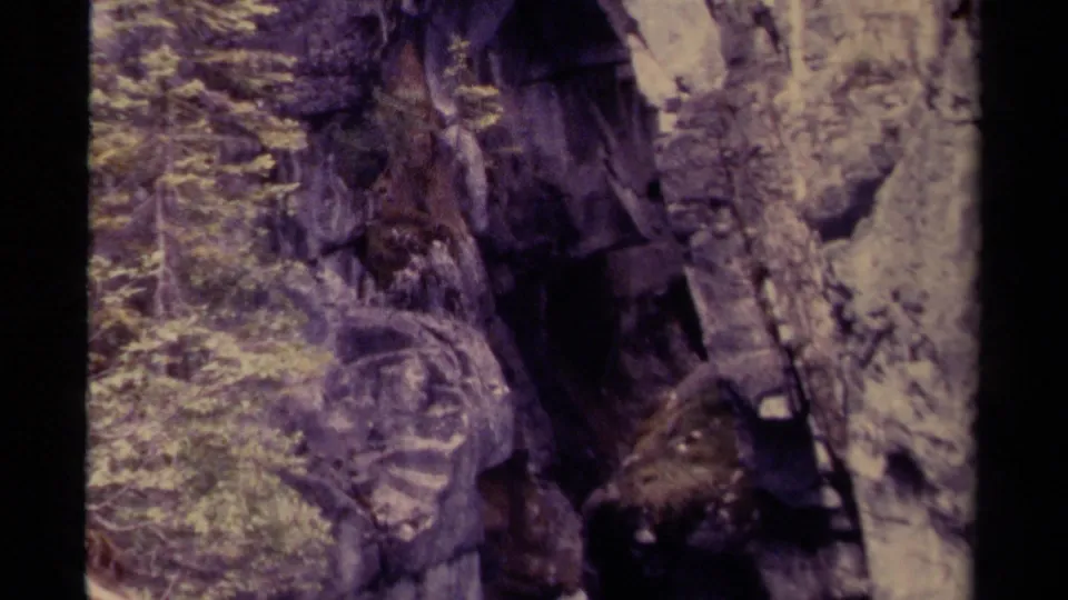 1979 Canadian home-movie frame with a dark cave-like opening, textured cliff wall, and evergreen growth nearby.