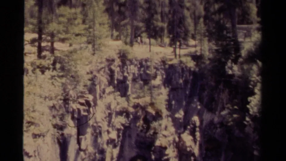 8mm home-movie frame from Canada showing a rugged cliff below evergreen trees, partly framed by dark foreground shapes.
