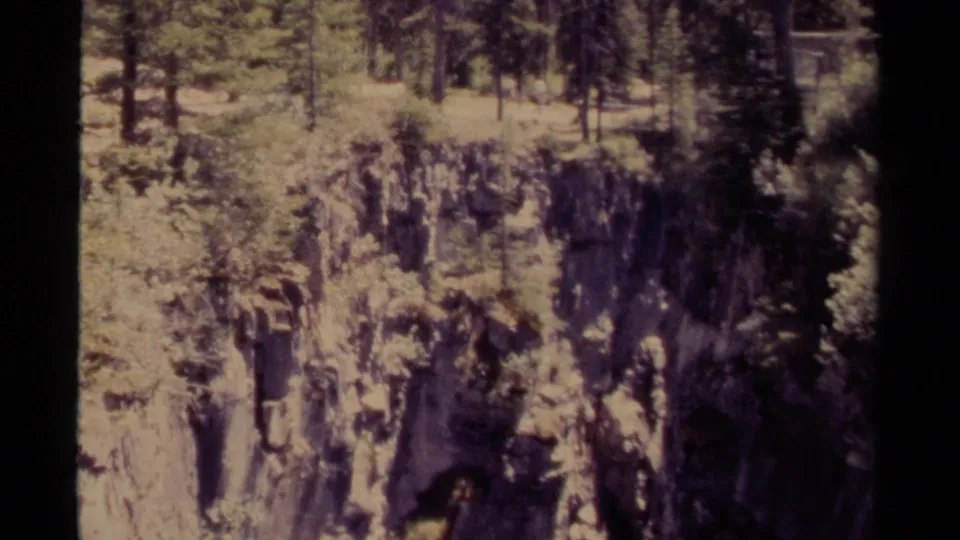 1979 Canada 8mm frame showing a rugged canyon wall, pale rock faces, sparse shrubs, and pine forest above.