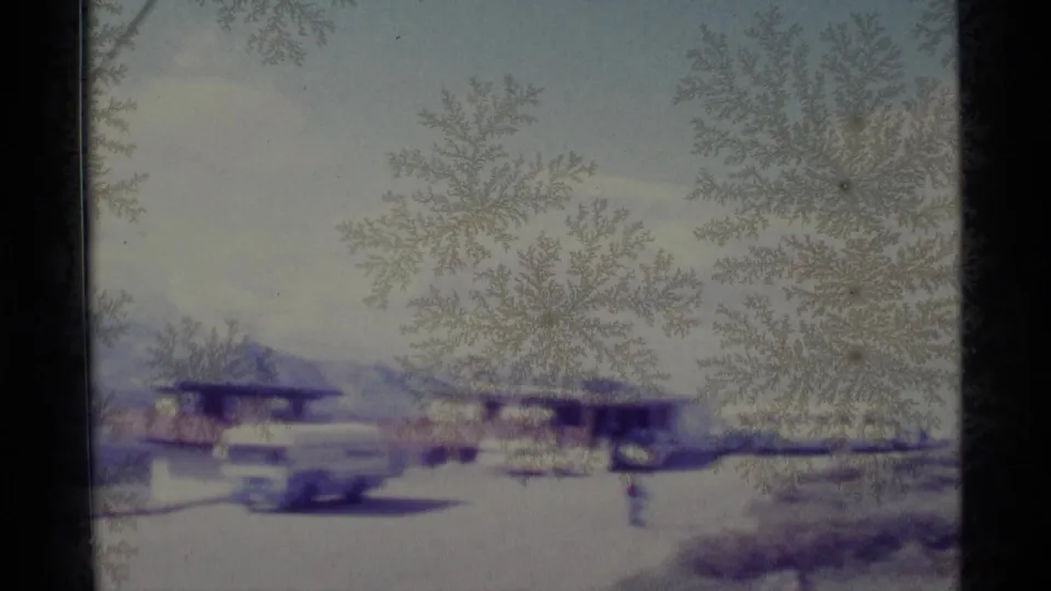 1977 Alaska home-movie frame looking through icy glass toward parked cars, low buildings, and distant mountains.