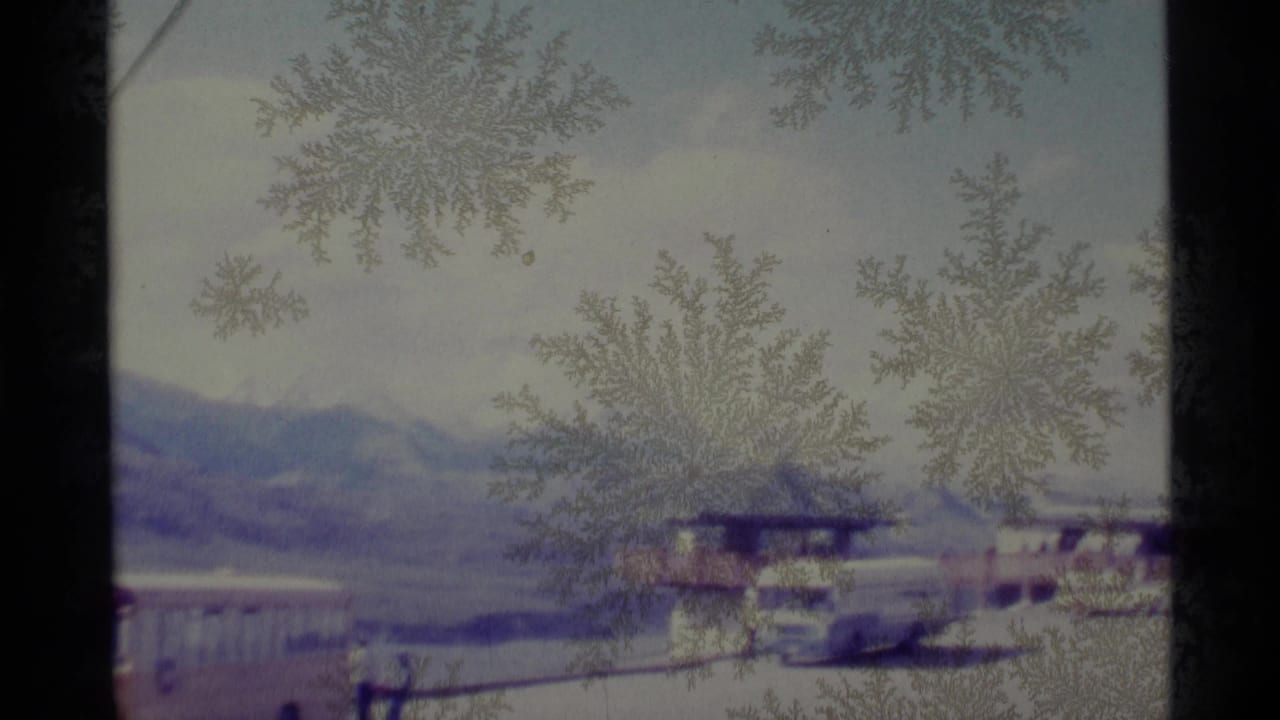 Frost patterns cover a 1977 Alaska roadside view with vehicles, structures, and a mountain range in the distance.