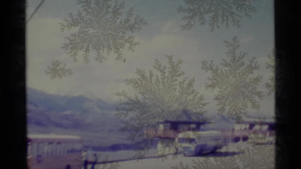 Frost patterns cover a 1977 Alaska roadside view with vehicles, structures, and a mountain range in the distance.