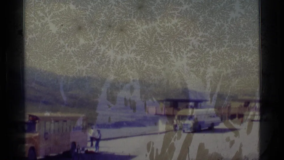 Visitors in a 1977 Alaska parking area walk near cars and a small building, partially obscured by frost on glass.