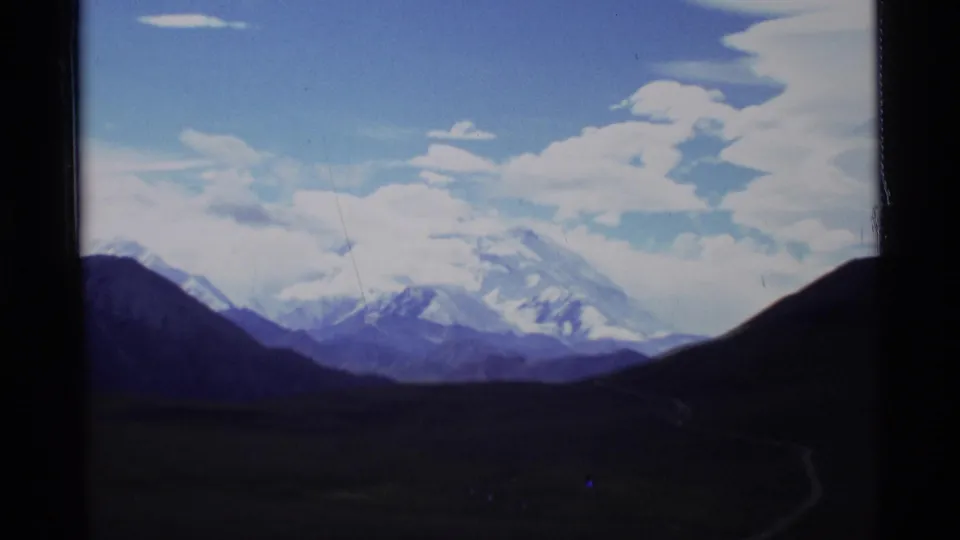 1977 Alaska scenic frame of snow-covered peaks, blue sky, scattered clouds, and a dark road or valley foreground.