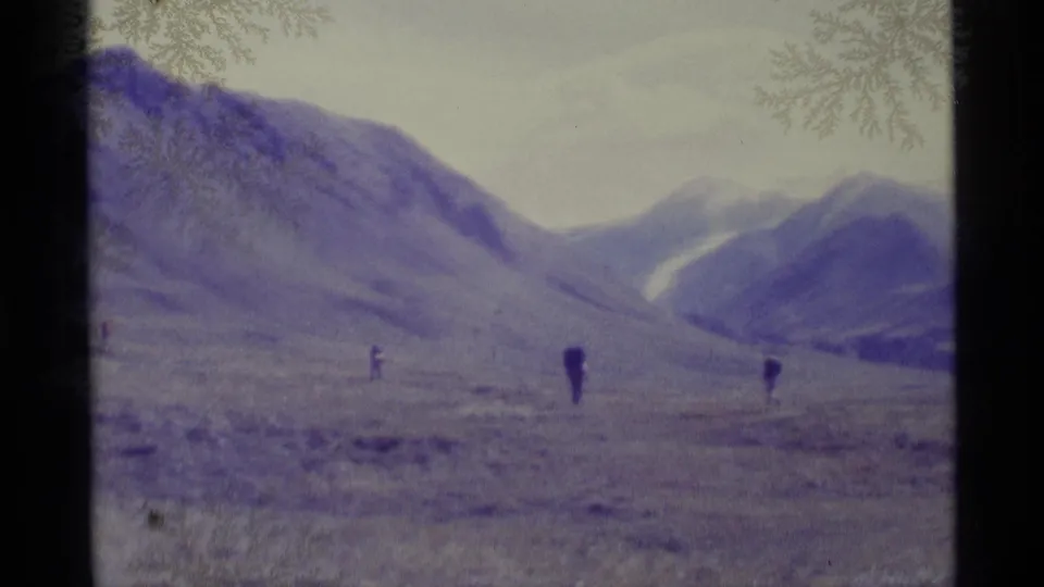 1977 Alaska 8mm frame shows several distant hikers on barren ground with mountains and a snow patch in the background.