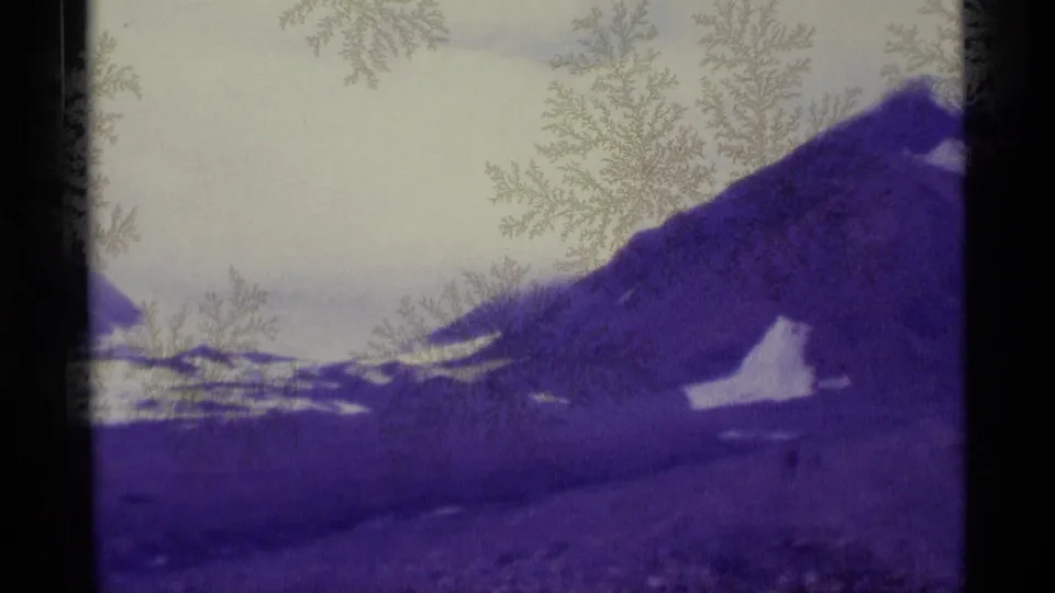1977 Alaska home-movie frame showing a purple mountain slope, snowy valley traces, pale sky, and frost-like film markings.