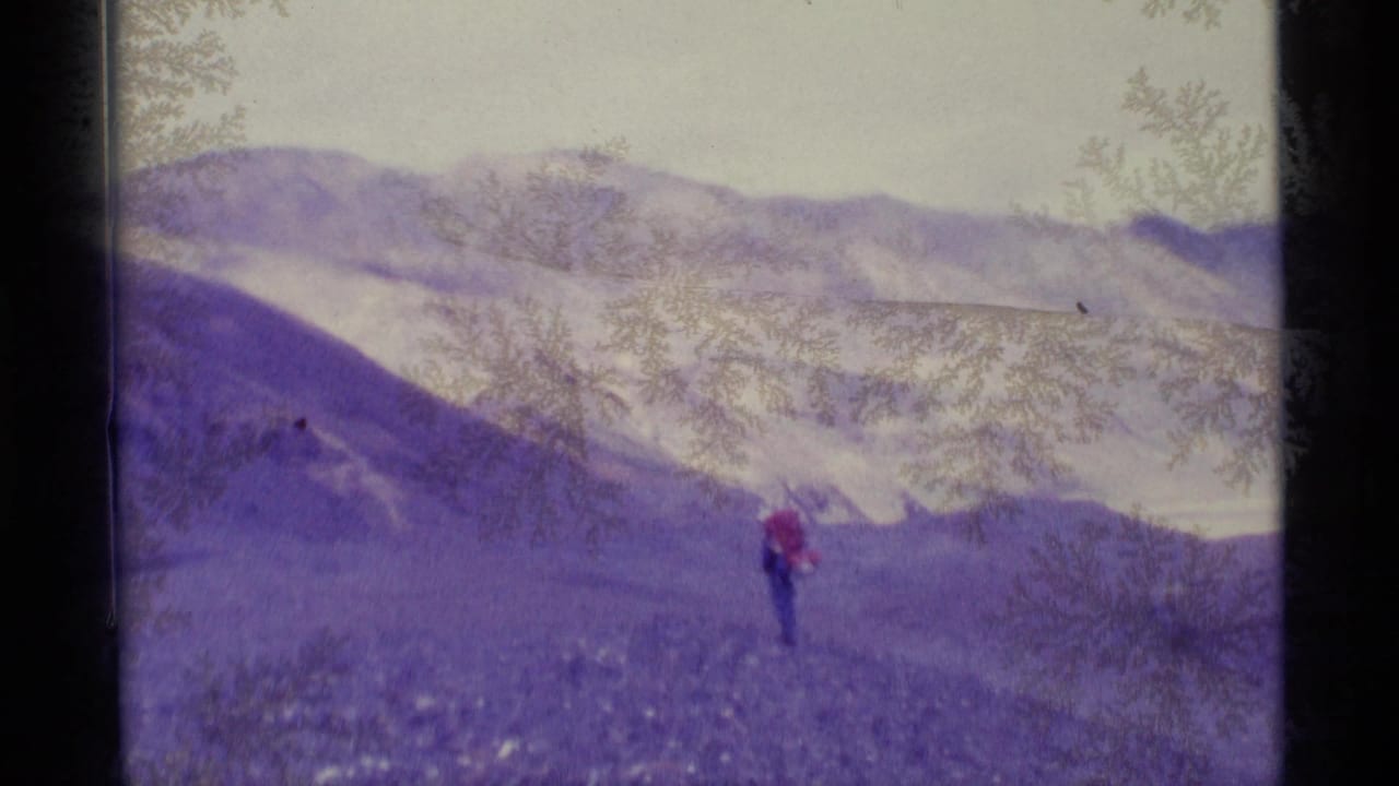 1977 Alaska home-movie frame with a lone distant hiker in red crossing rocky terrain beneath snow-marked ridgelines.