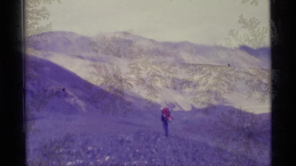1977 Alaska home-movie frame with a lone distant hiker in red crossing rocky terrain beneath snow-marked ridgelines.