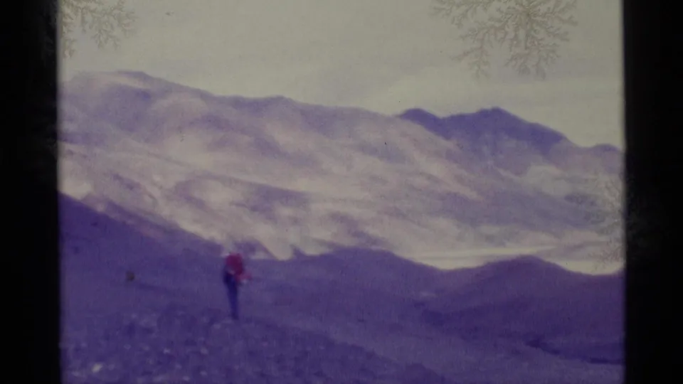 1977 Alaska 8mm frame showing a distant red-jacketed hiker on rough ground before pale mountain ridges.