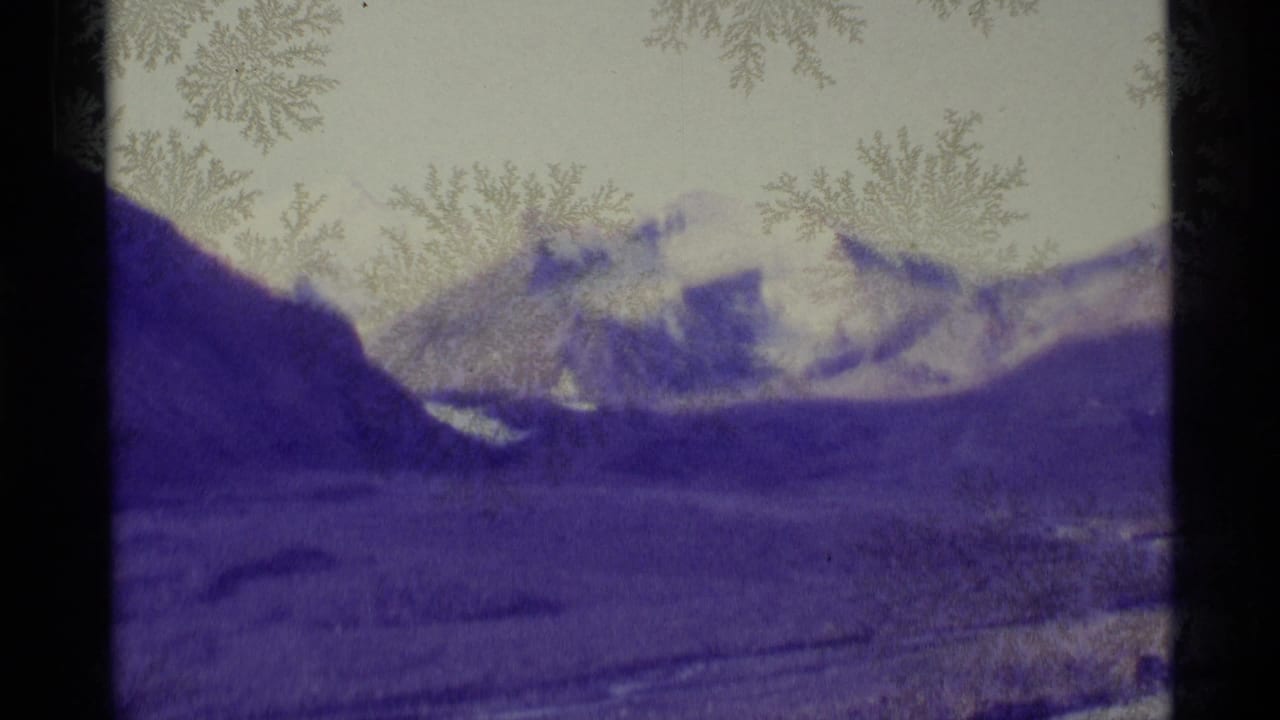 1977 Alaska 8mm frame of distant snow-capped mountains and a valley, strongly purple-tinted with visible film edge artifacts.