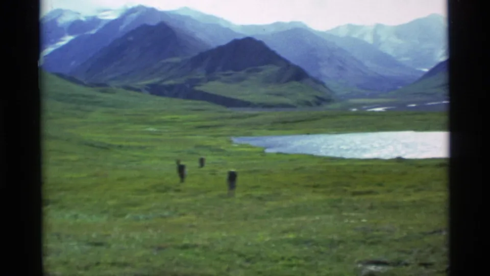 1977 Alaska 8mm frame showing three distant hikers crossing green tundra beside a small lake and mountain range.
