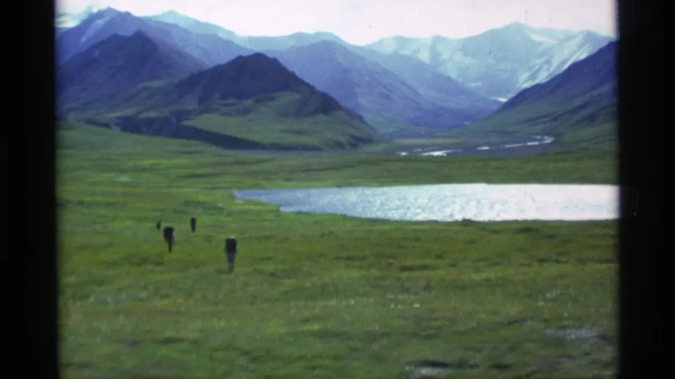 A 1977 8mm frame shows tiny figures crossing a meadow before a shimmering lake and layered mountain ridges.