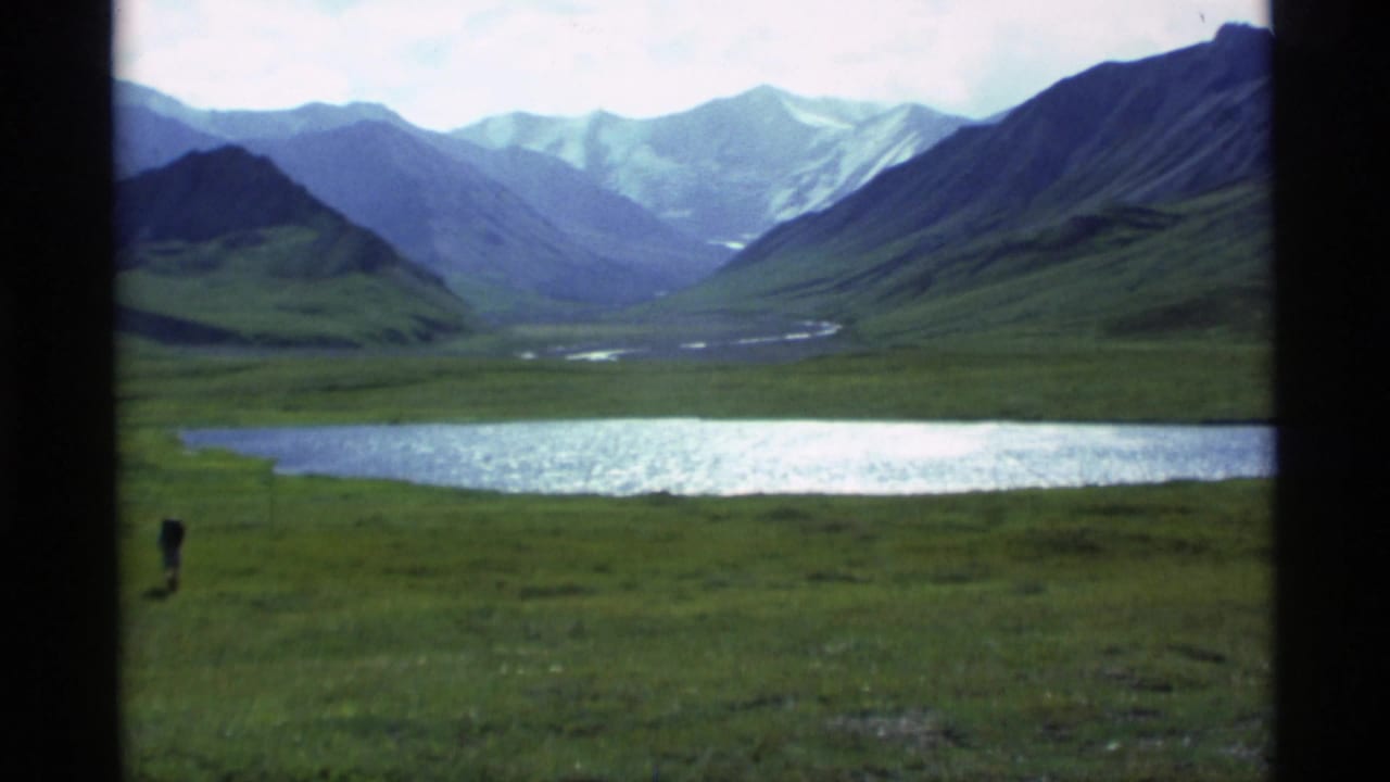 The pan continues across meadow and lake toward a mountain valley, with one small human figure visible in the grass.