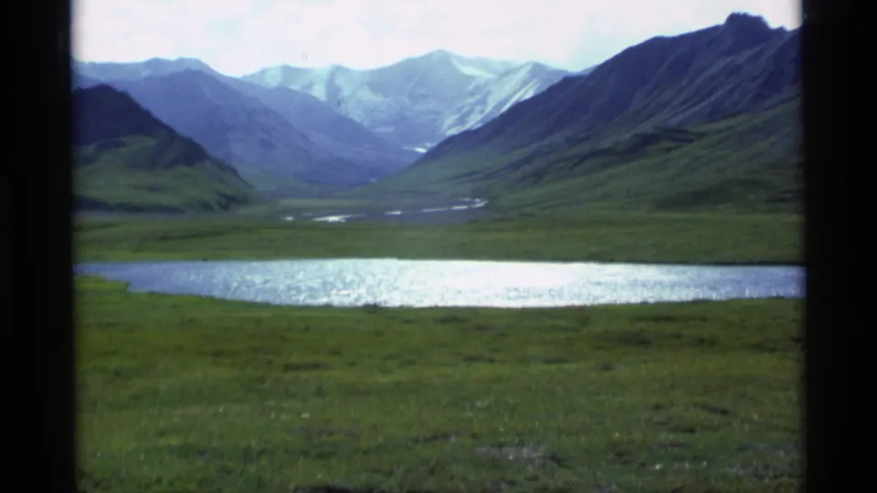 A soft 8mm frame shows a reflective lake stretching across the foreground beneath a deep Alaskan mountain valley.