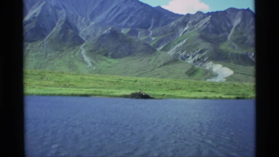 After the cut, the frame shows closer blue water, a grassy shoreline, and rugged green-gray mountain slopes.