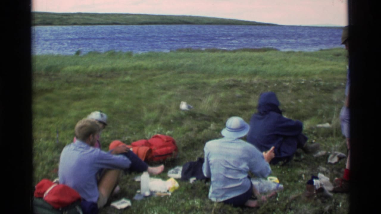 1977 Alaska home-movie frame showing tourists seated on a grassy shoreline beside a blue lake, with red bags and picnic items nearby.