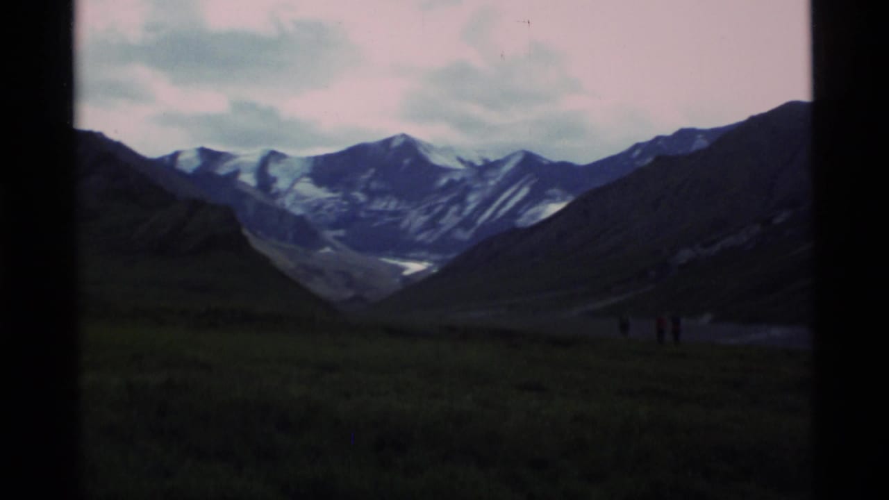 Small hikers cross a green alpine valley with snow-capped mountains in the distance.