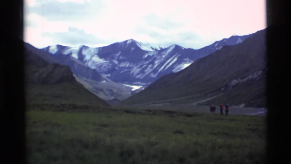 1977 Alaska home-movie view of hikers near the right edge of a green valley framed by snowy peaks and dark ridges.